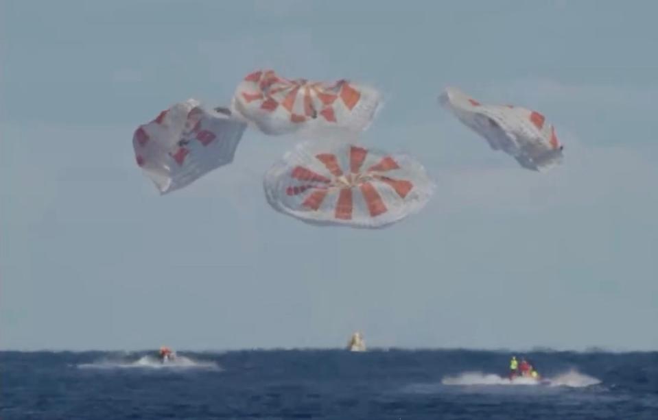 a cone-shaped spacecraft crashing into the ocean with parachutes deflating above it. Boats in the water race toward the spacecraft