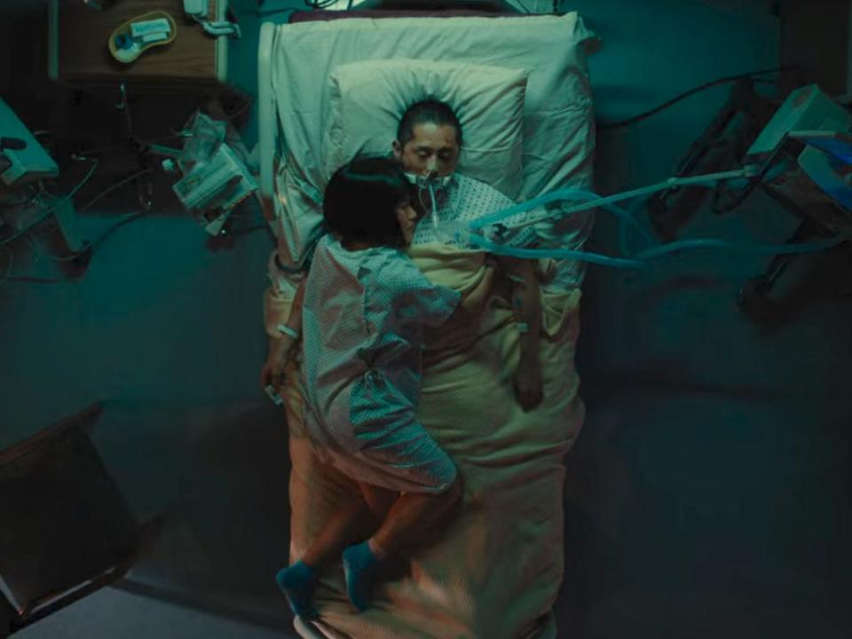 danny lying in a hospital bed in beef, with amy draped over him and resting her head on his chest. danny is unconscious and hooked up to a variety of machines, including a breathing tube, and the top of his hospital bed