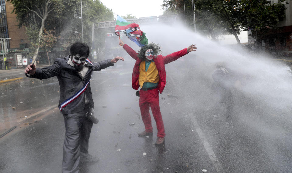 Men dressed as clowns, one dressed as the the movie character “The Joker” flying a Mapuche indigenous flag, are sprayed by a police water cannon during an anti-government protest in Santiago, Chile, Monday, Nov. 4, 2019. Chile has been facing weeks of unrest, triggered by a relatively minor increase in subway fares. The protests have shaken a nation noted for economic stability over the past decades, which has seen steadily declining poverty despite persistent high rates of inequality. (AP Photo/Esteban Felix)