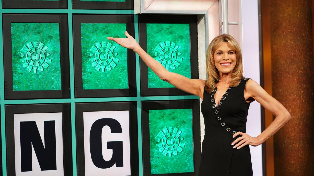 vanna white, wearing a black dress, gesturing toward the wheel of fortune puzzle