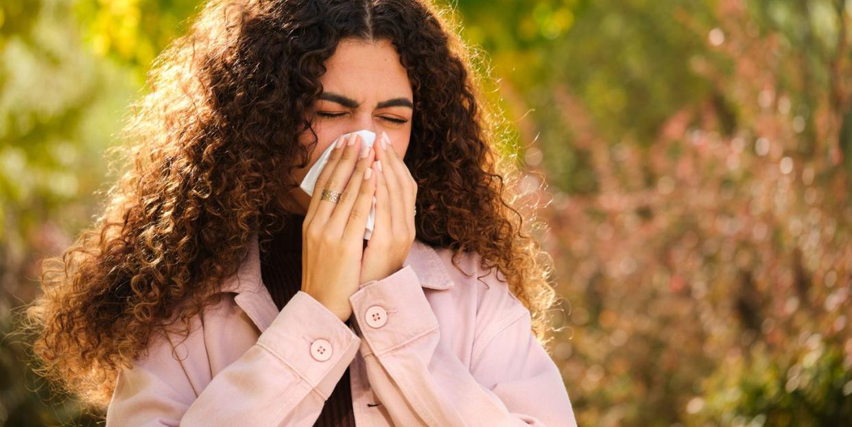 young woman with curly hair blowing your nose in a tissue on tree lined street