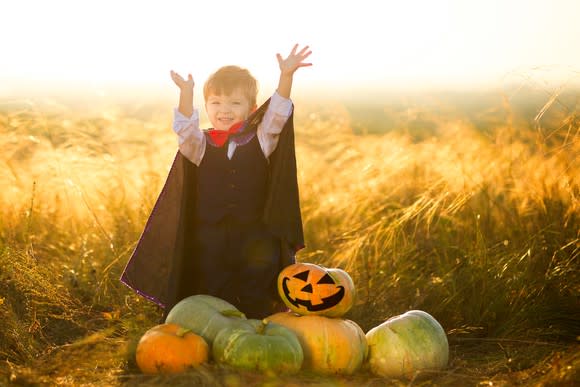 A child in a Dracula costume standing behind a pile of pumpkins in a field.