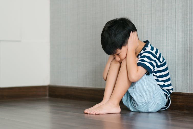 A little boy in a stripey T-shirt sits on the floor with his hands over his ears.