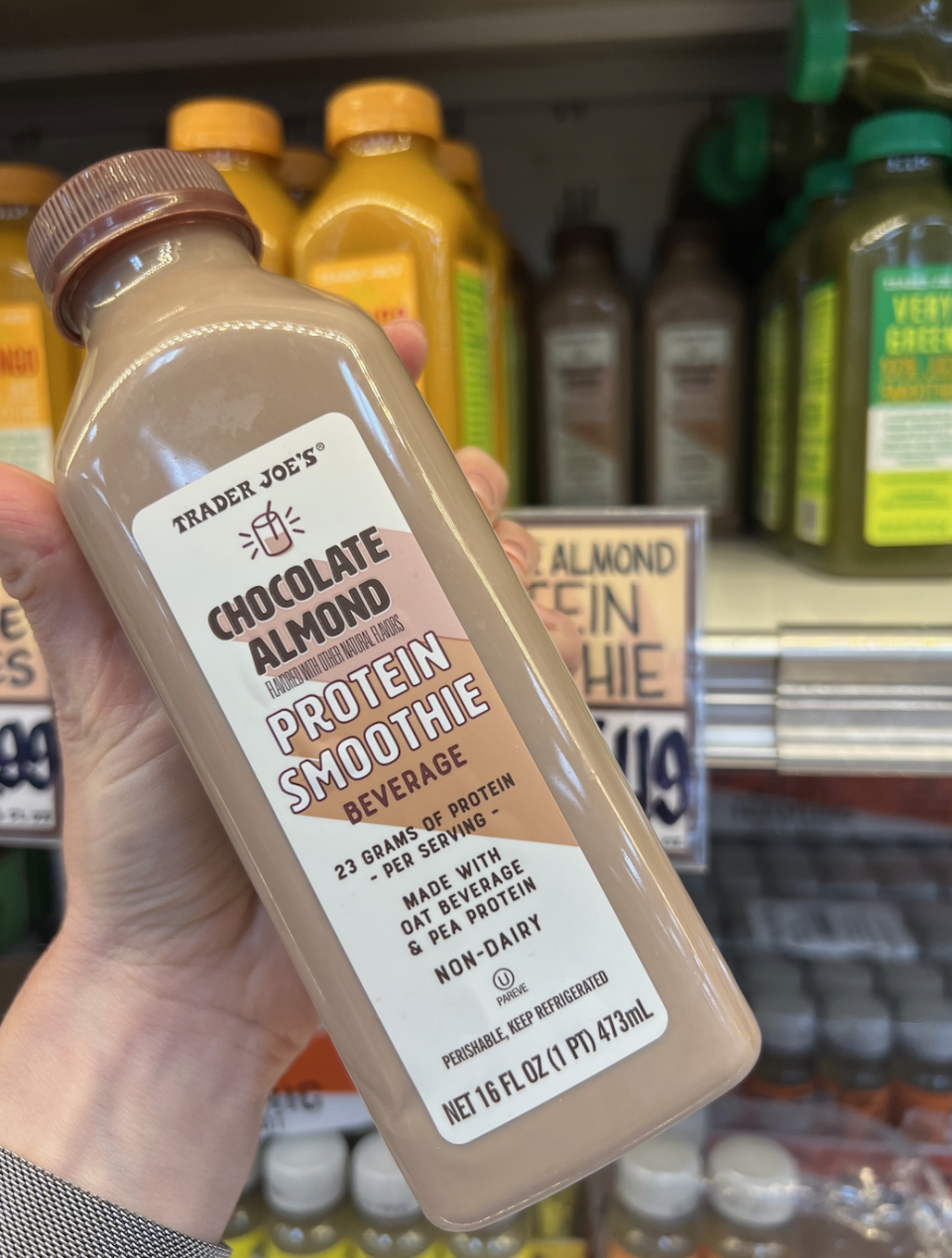 Hand holding a bottle of Trader Joe's Chocolate Almond Protein Smoothie beverage in front of store shelves stocked with similar drinks