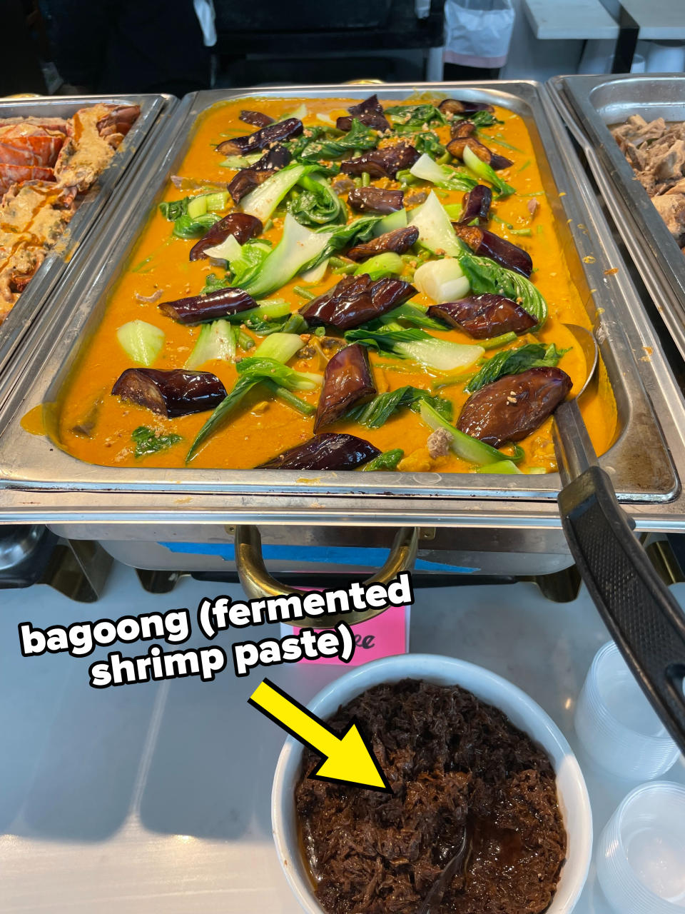 Buffet tray filled with a dish labeled "Kare Kare" featuring vegetables, eggplant, and a yellow sauce with a serving utensil. Bowl of dark shredded meat nearby