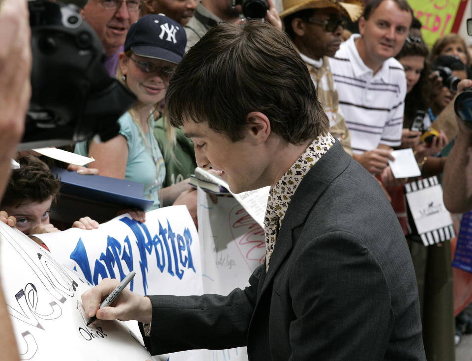 Daniel Radcliffe signing autographs, wearing a jacket and patterned scarf, with fans and cameras around him
