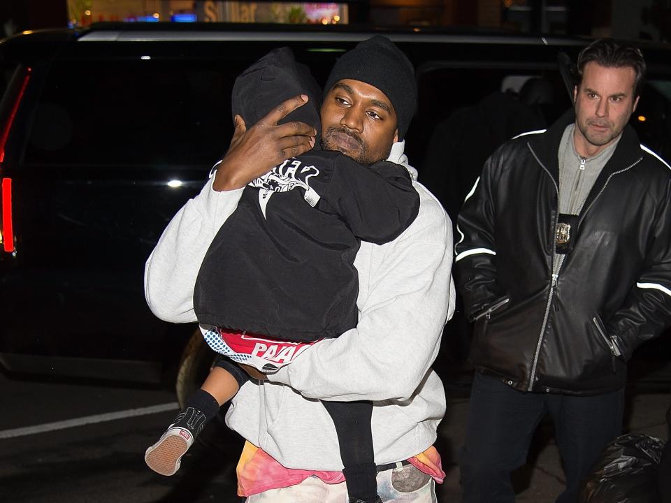 steve stanulis in the background of a photo of ye, cradling his daughter north west to his chest, and walking on the street. north is very young in this photo, her face away from the camera, and stanulis stands a few paces behind ye, seemingly looking in front of him