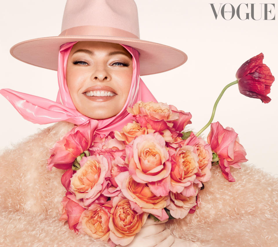 Spread from the Vogue feature with Linda smiling, wearing a hat and attached scarf, and holding a bouquet of flowers