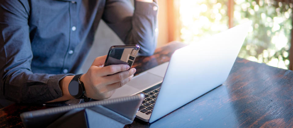 Male hand holding smartphone. Businessman using laptop computer and digital tablet while working in the cafe. (Getty Images)