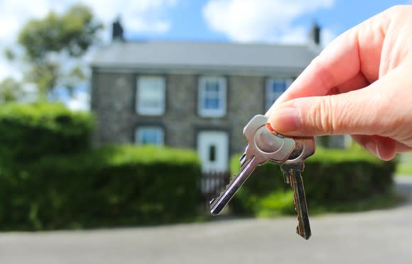 Hand holding two keys in the foreground of a two-story home.