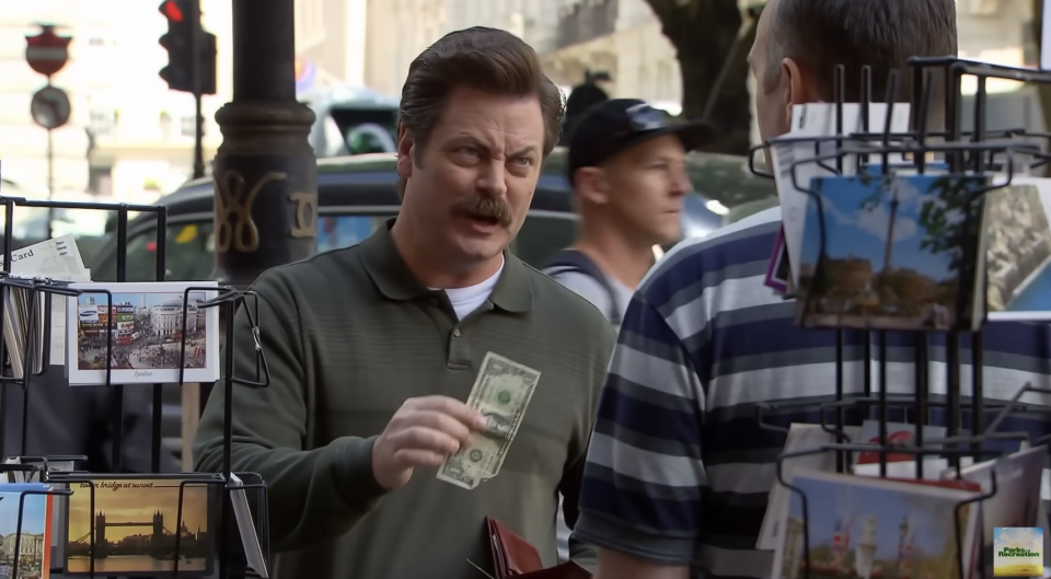 Nick Offerman in Parks and Rec holds up a dollar bill, talking to another man in a striped shirt at a postcard stand on a city street