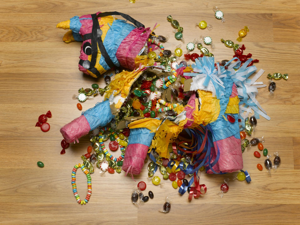 A broken piñata lies on the floor with various candies and small toys scattered around it. The festive piñata appears to be in the shape of a donkey