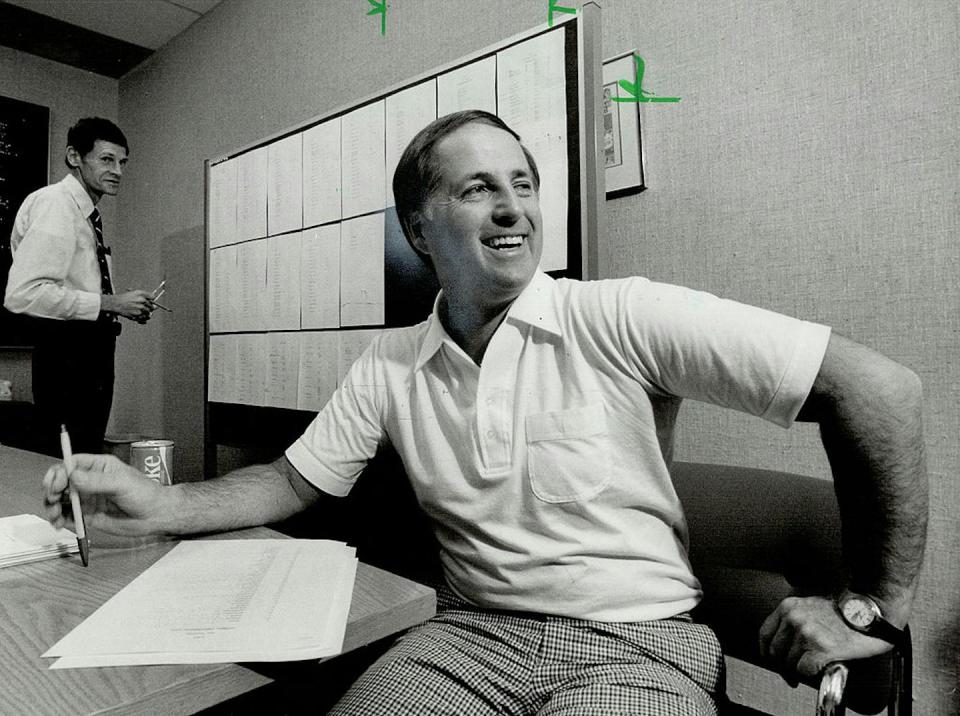 Black and white photo of smiling man seated at a table. Behind him is another man standing next to a board with sheets of paper affixed to it.