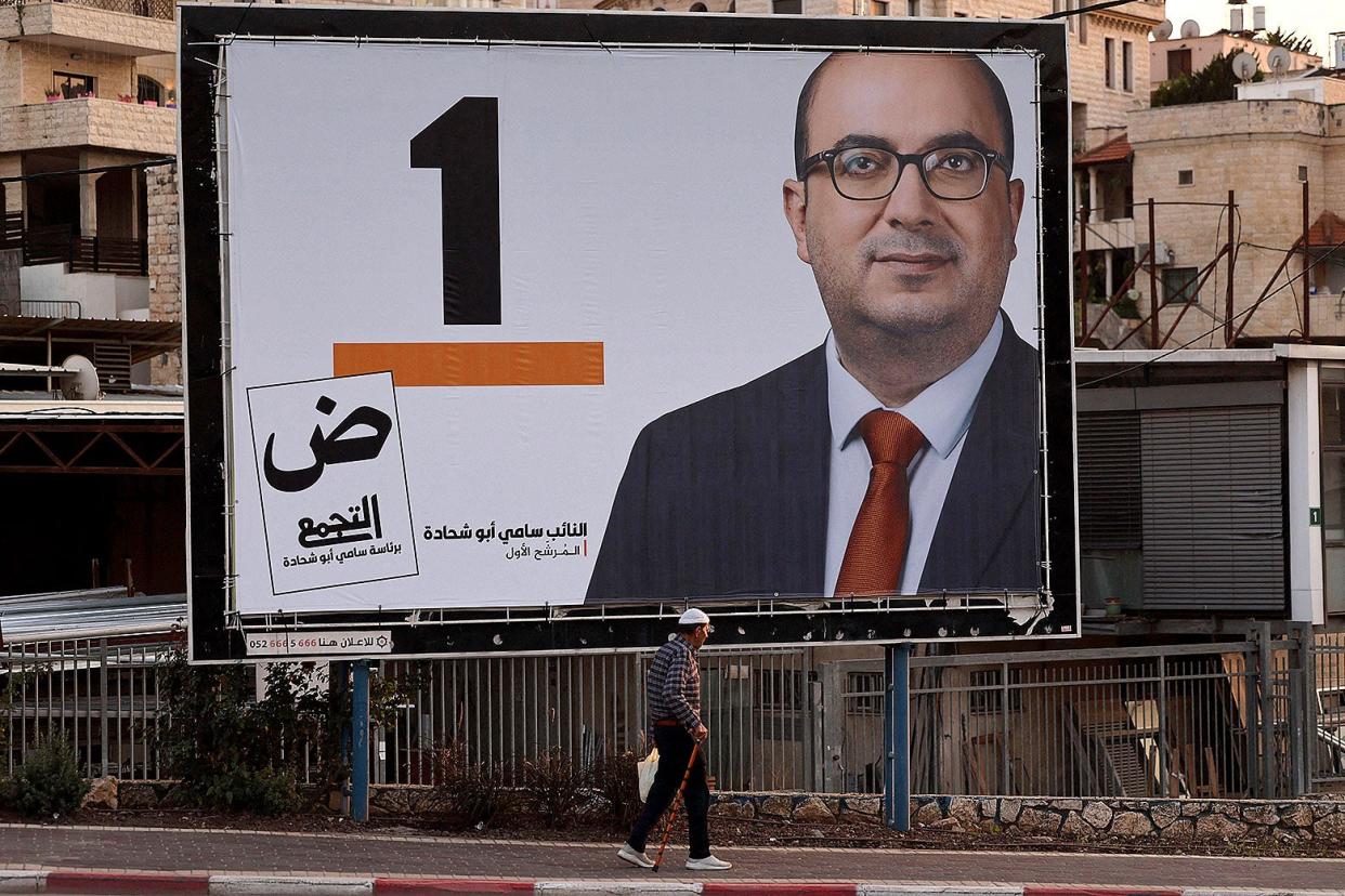 An electoral billboard of Sami Abu Shehadeh displays a middle-aged man in glasses wearing a red tie as people walk on the sidewalk underneath. 