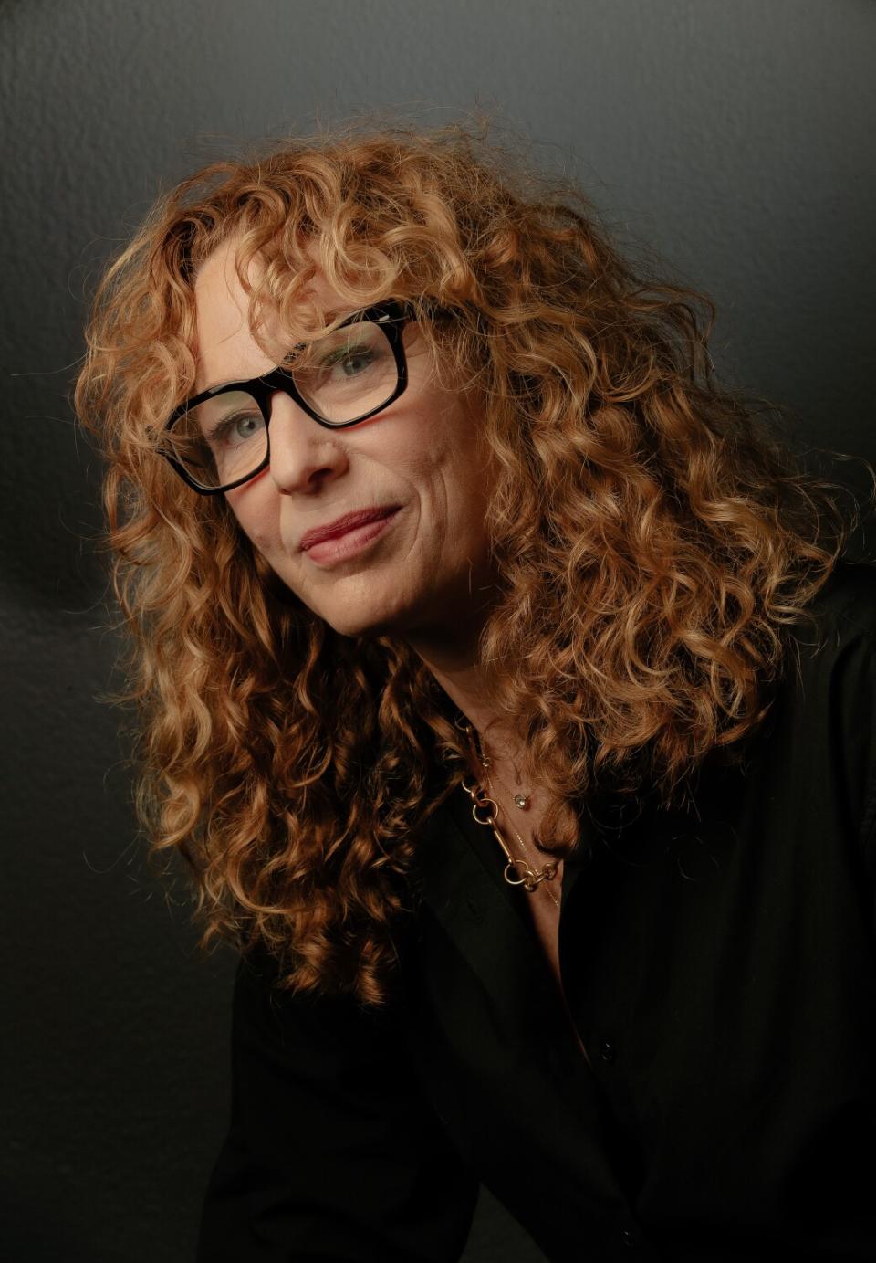 A portrait of a woman with red curly hair and dark-framed glasses.