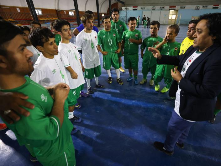 Gente pequeña con ropa de fútbol escucha en un polideportivo a un hombre con una chaqueta de estatura similar.