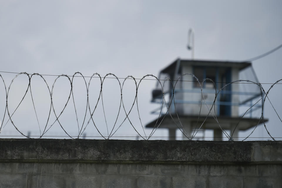 Barbed wire atop a prison wall with a guard tower in the background.