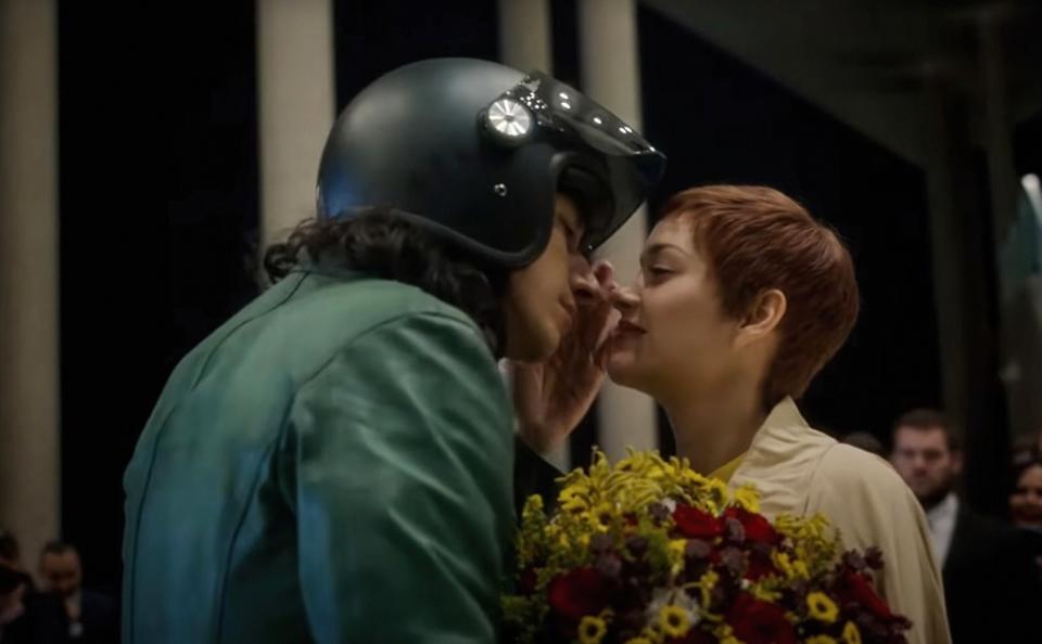 Marion Cotillard holding flower prepares to kiss Adam Driver in a motorcycle helmet