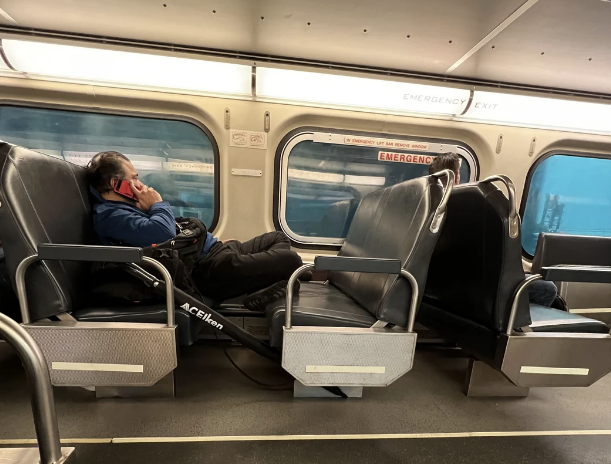 A person is lying across train seats with their head resting on a pillow and wearing headphones. Another passenger is partially visible in the background