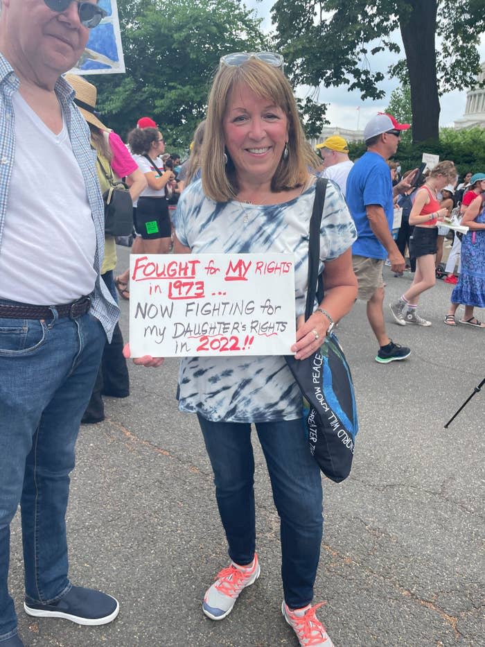A woman in a tie-dye shirt holds up a sign reading "Fought for my rights in 1973... now fighting for my daughter's rights in 2022!!"