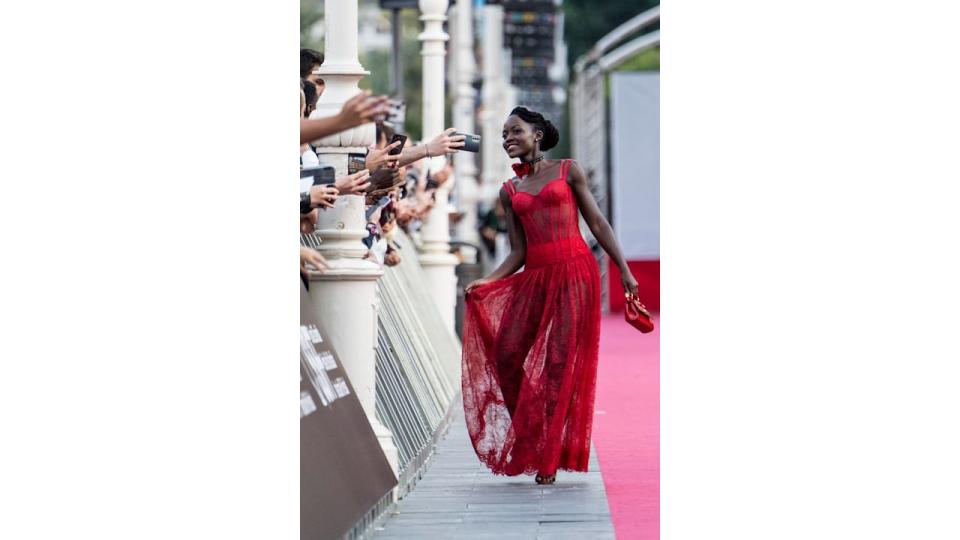 Lupita Nyong'o, dressed in a red lace gown, walks gracefully on the red carpet while interacting with fans. She holds her flowing skirt with one hand, revealing its semi-sheer design, while her other hand clutches a red handbag.