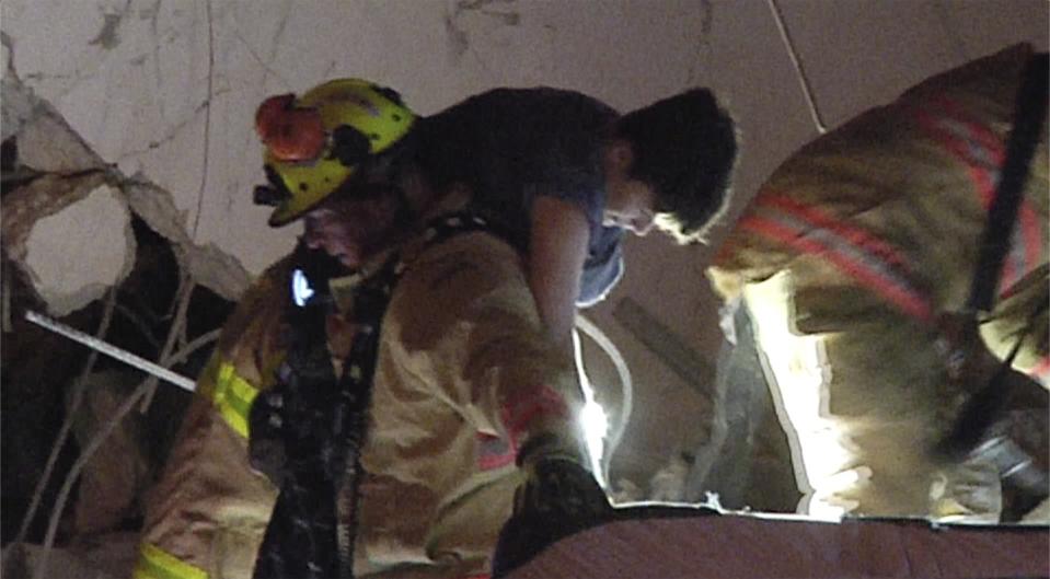 A firefighter carries a person. A gaping, broken wall is seen in the background.