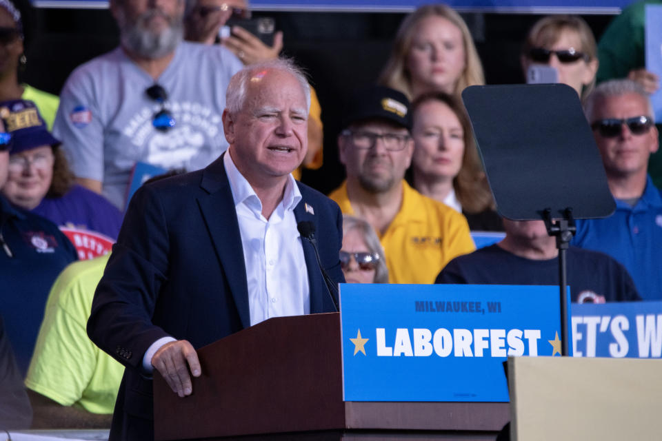 Governor Tim Walz speaks at a podium with a "Milwaukee, WI Laborfest" sign, surrounded by attendees in casual clothing, some wearing caps and sunglasses