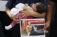 In this Jan. 12, 2019 photo, firefighter Fatima Olmedo holds her two-month-old baby Samara as her husband holds a calendar page with a photo Olmedo posing nude while pregnant, in Asuncion, Paraguay. Olmedo was first to pose naked for the calendar, holding a rolled-up hose in her hands that barely covered her pregnant belly. (AP Photo/Jorge Saenz)
