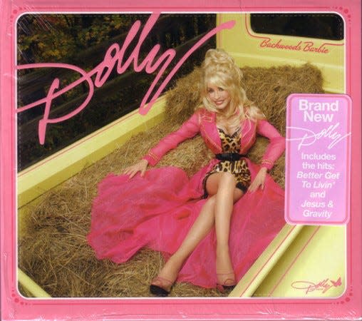 Dolly Parton lounging on hay stacks in the back of a yellow pickup truck wearing a leopard print minidress belted with a long, sheer pink overcoat/dress.