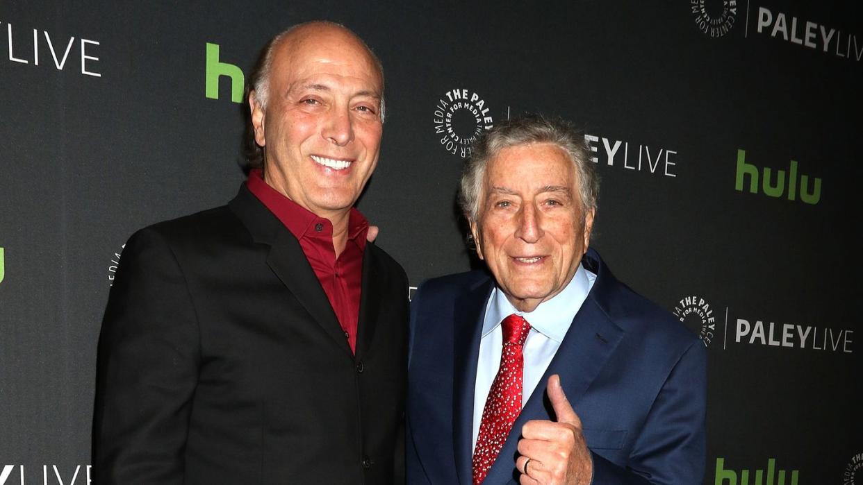 danny bennett and tony bennett embrace and pose for a photo while standing in front of a black backdrop with logos, both men smile and tony flashes a thumbs up, danny wears a black suit jacket and red collared shirt, tony wears a navy suit jacket, light blue collared shirt and red tie with a white pattern on it