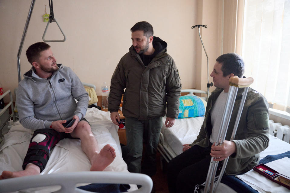 Zelensky stands between two hospital beds, on which sit two men, one in a leg cast and the other holding crutches.