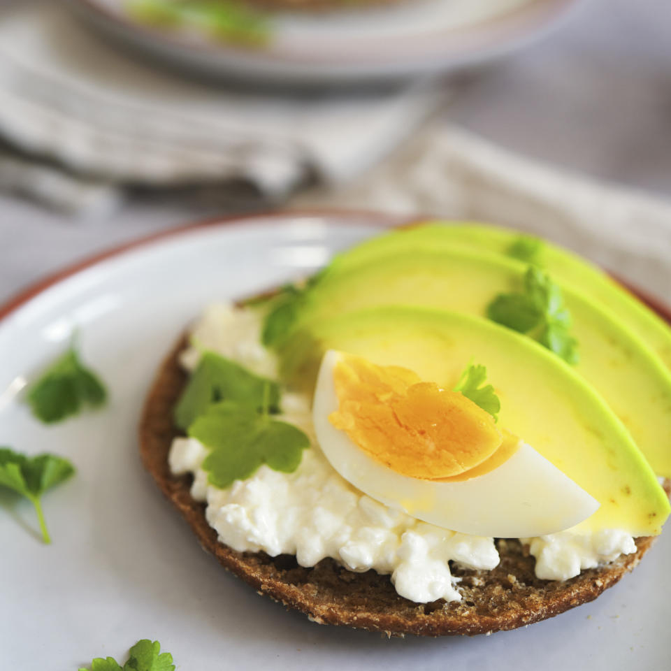 A rye bread slice topped with cottage cheese, avocado slices, a boiled egg half, and parsley leaves on a white plate