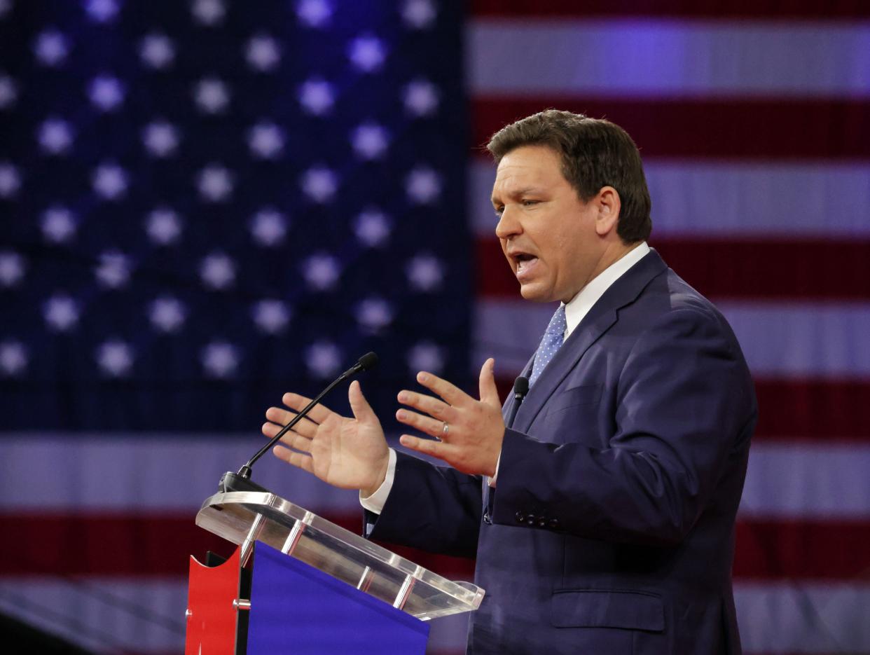 Ron DeSantis speaks into a microphone in front of a large American flag image.