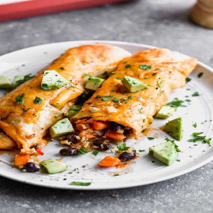 Two enchiladas on a plate topped with red sauce, diced avocado, and fresh herbs