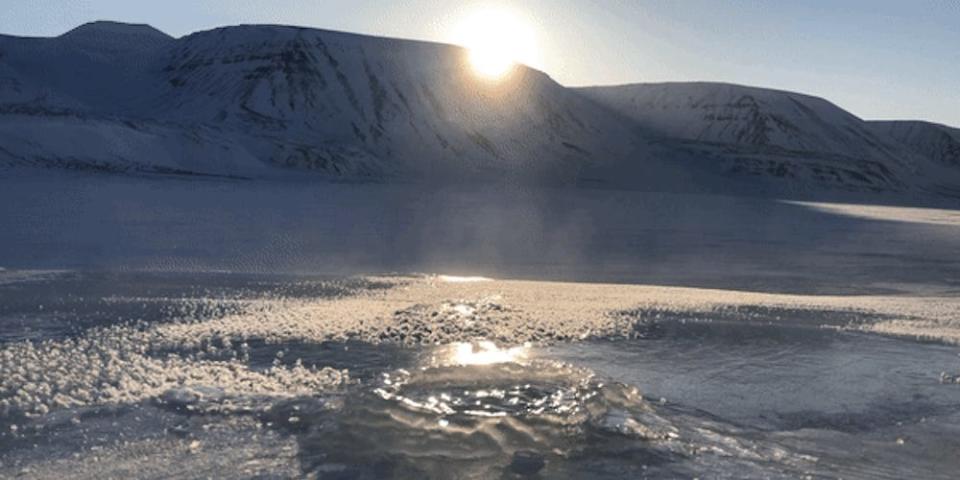 Water bubbles with methane as it pierces through a sheet of ice. In the background are snow covered mountains of Svalbard.