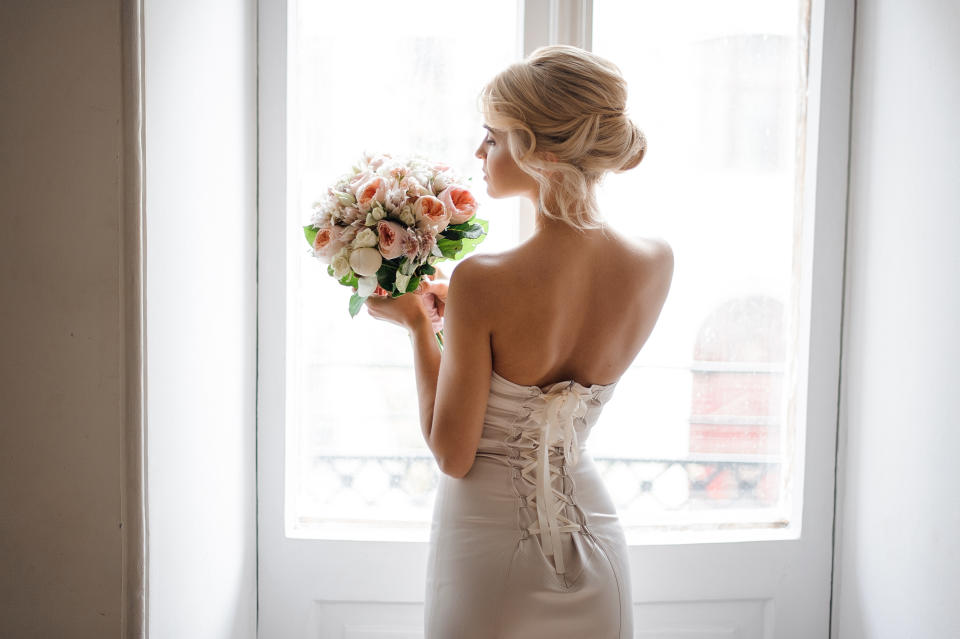 Back view of the elegant blonde bride dressed in a white dress holding a wedding bouquet on the background of window.