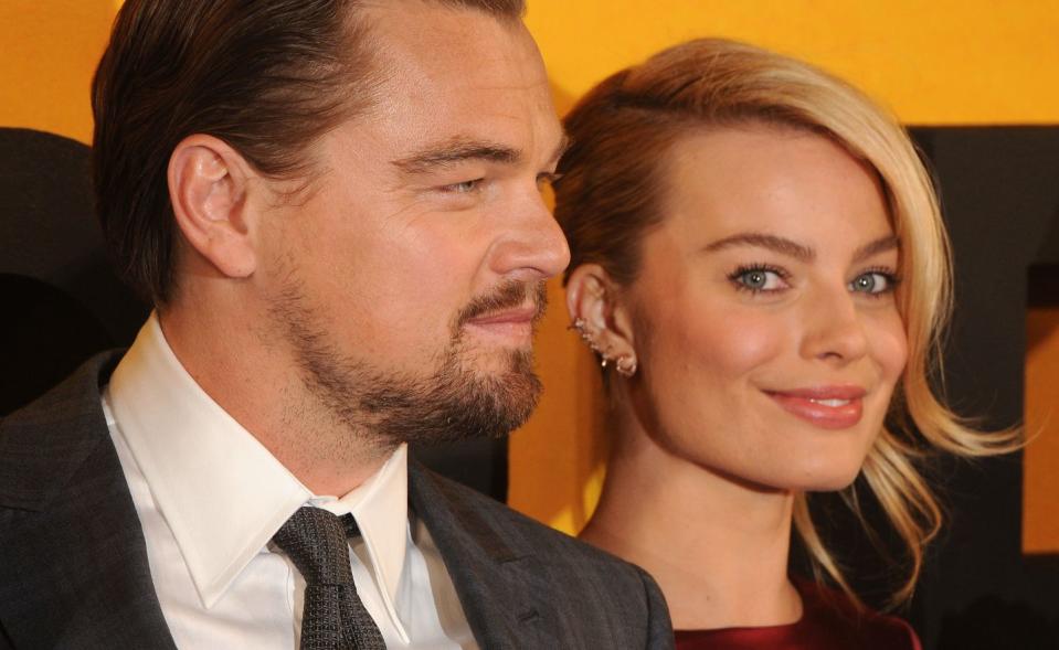 margot robbie smiles at the camera, she wears a red dress and several earrings, leonardo dicaprio is to her left and stares straight ahead in profile