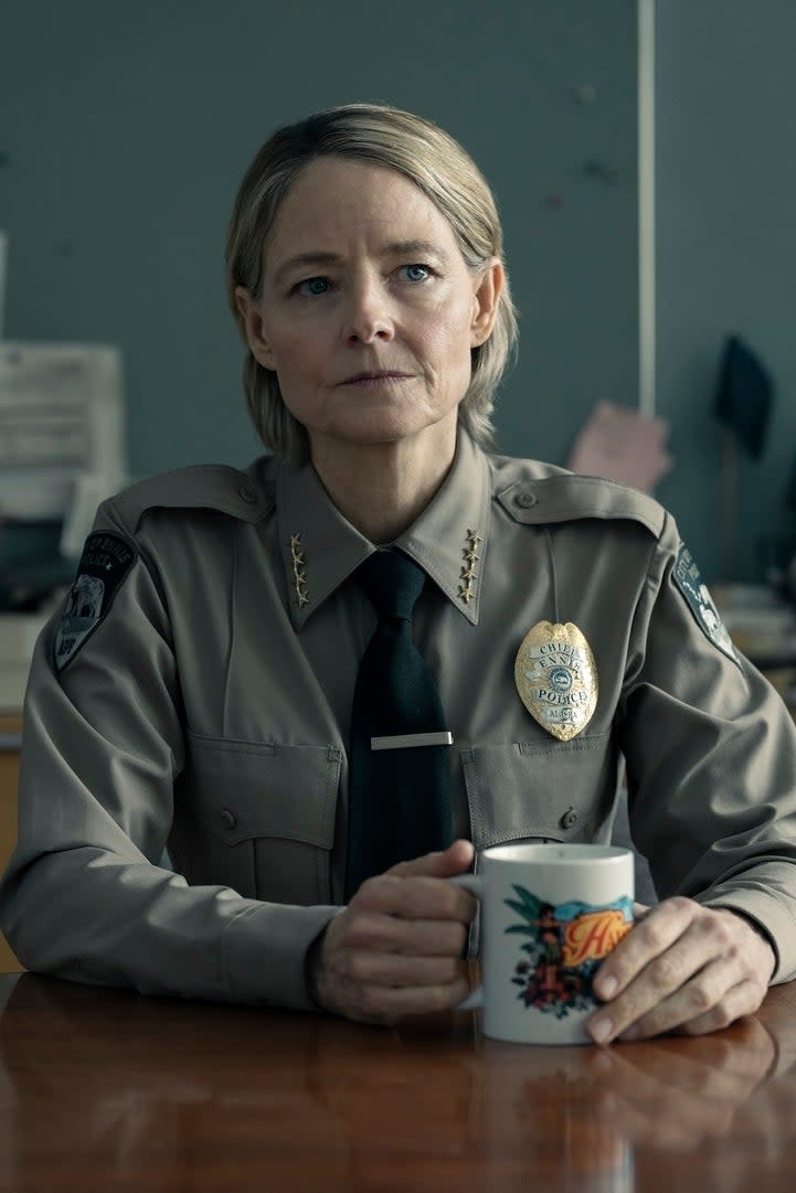 Jodie Foster as a uniformed sheriff, sitting at a wooden table holding a coffee mug, in a formal office setting with file folders and books behind her