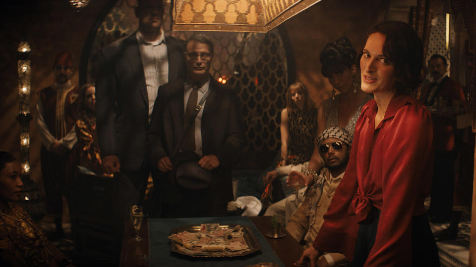 Still from the movie Indiana Jones and the Dial of Destiny. Here we see a pile of money on a silver platter on top of a table inside a bazaar-style room. Surrounding the table are a number of suspicious-looking men and woman.