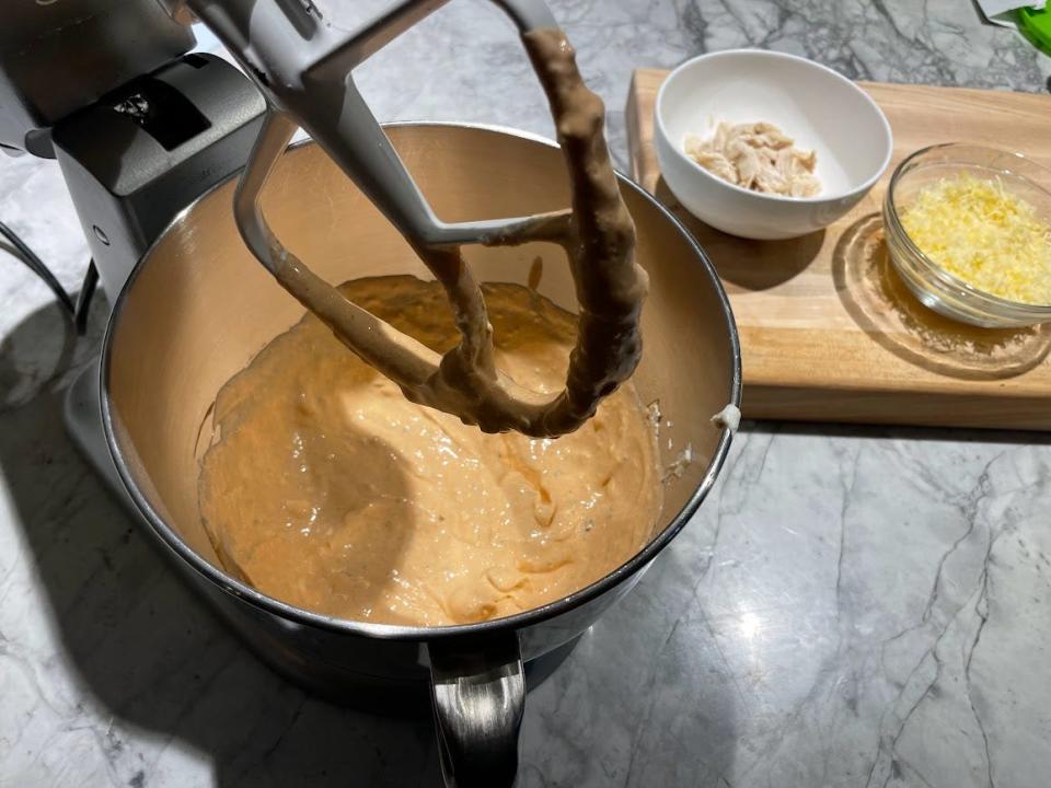 A mixer with the bowl filled with orangey partially made Buffalo-chicken dip. Bowls of shredded cheese and chicken on a cutting board are in the background