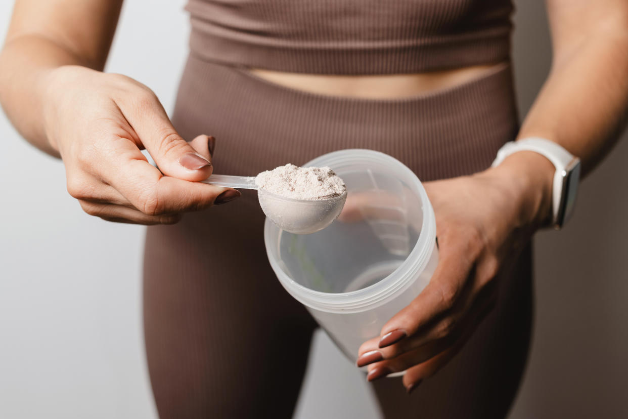 Athletic woman in sportswear with measuring spoon in her hand puts a portion of whey protein powder into a shaker, making protein drink, cropped image.
