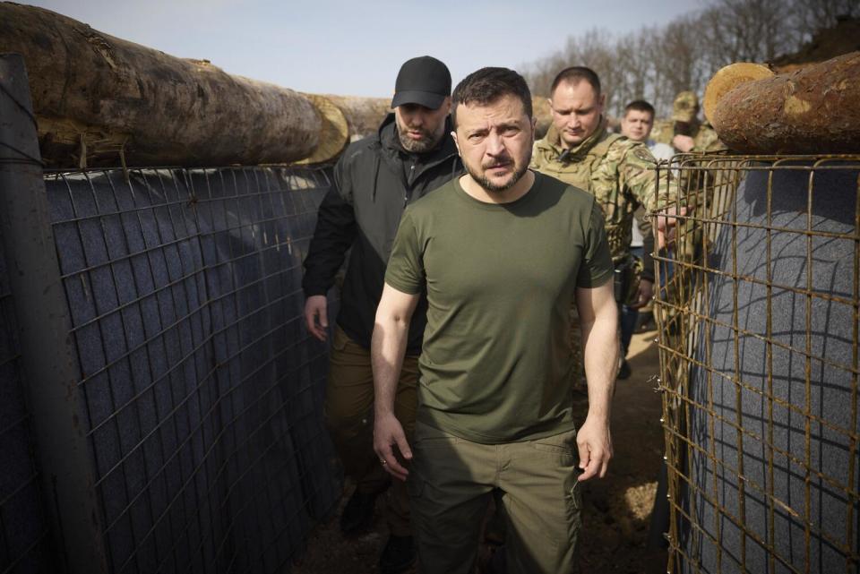 A man with dark close-cropped hair and beard, in an olive green shirt and pants, stands in a trench with other people