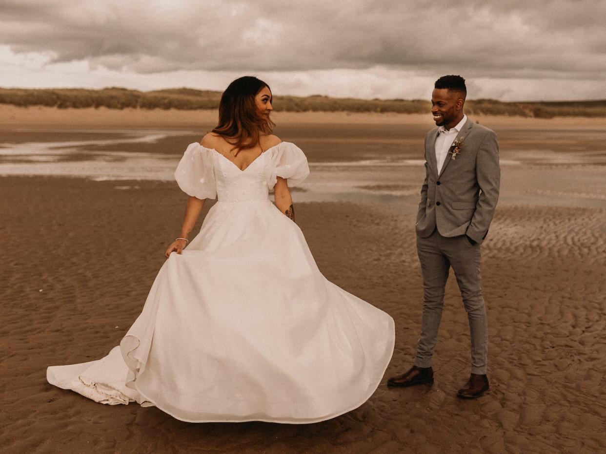 A bride and groom look at each other on a beach in their wedding attire.