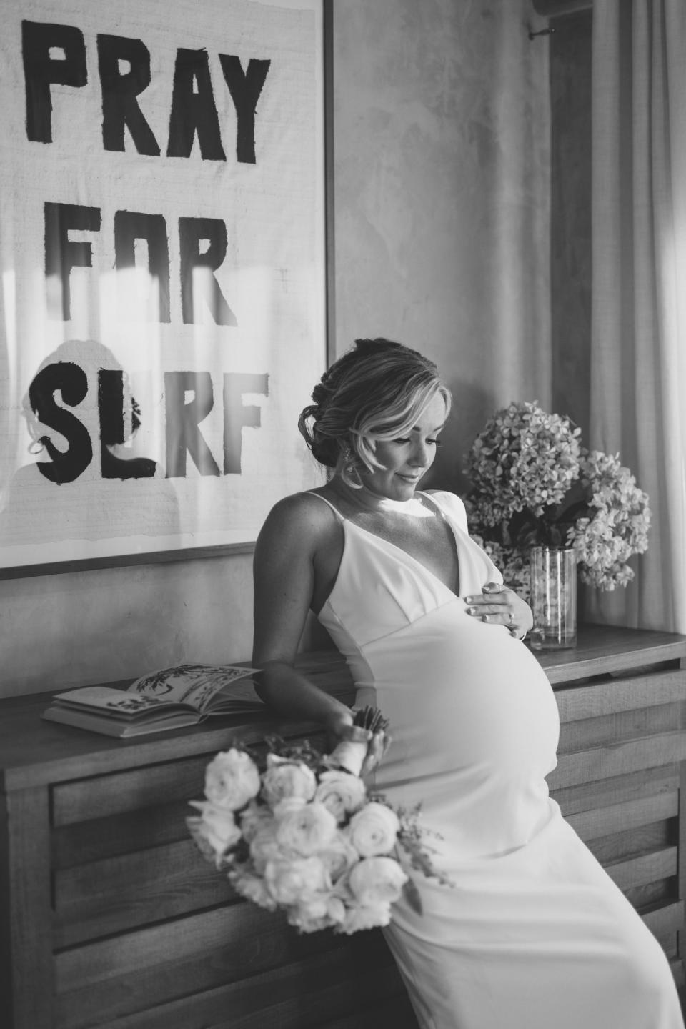 A bride leans back against a bookshelf. One of her hands rests on her pregnant belly, and she holds a bouquet in her other hand.