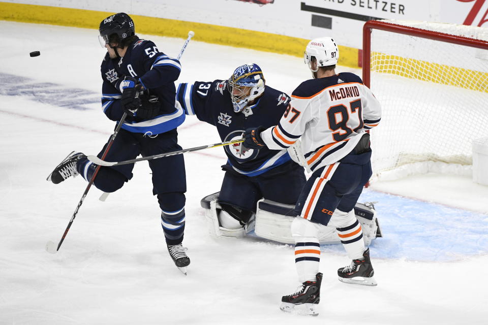 A shot by the Edmonton Oilers goes wide of the net past Winnipeg Jets' Mark Scheifele (55) and goaltender Connor Hellebuyck (37) as Oilers' Connor McDavid (97) looks for the deflection during first-period NHL hockey Stanley Cup playoff action in Winnipeg, Manitoba, Sunday, May 23, 2021. (Fred Greenslade/The Canadian Press via AP)