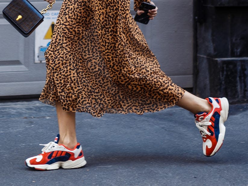 woman walking down a street wearing a long cheetah print dress and trendy red sneakers