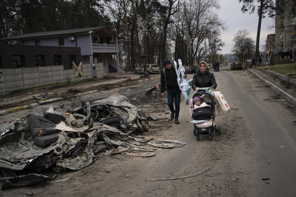 A family walks pass a car crushed by a Russian tank in Bucha, in the outskirts of Kyiv, Ukraine, Tuesday, April 5, 2022. Ukraine's president planned to address the U.N.'s most powerful body on Tuesday after even more grisly evidence emerged of civilian massacres in areas that Russian forces recently withdrew from. (AP Photo/Rodrigo Abd)