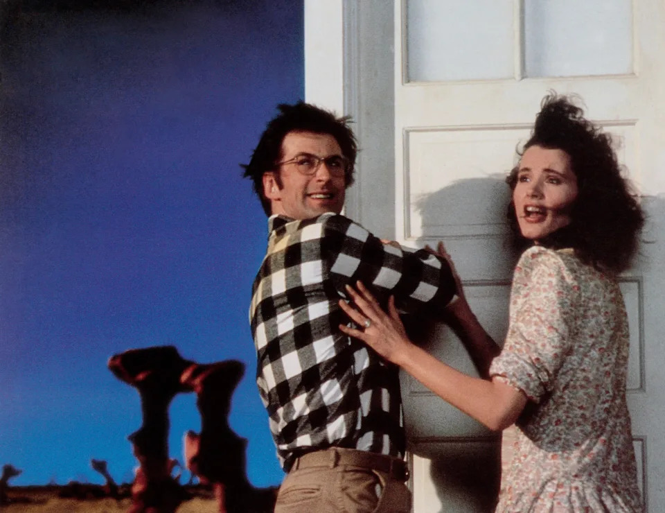 Jeff Goldblum and Geena Davis in a dramatic scene, holding a door closed against an unseen force, with a surreal landscape in the background