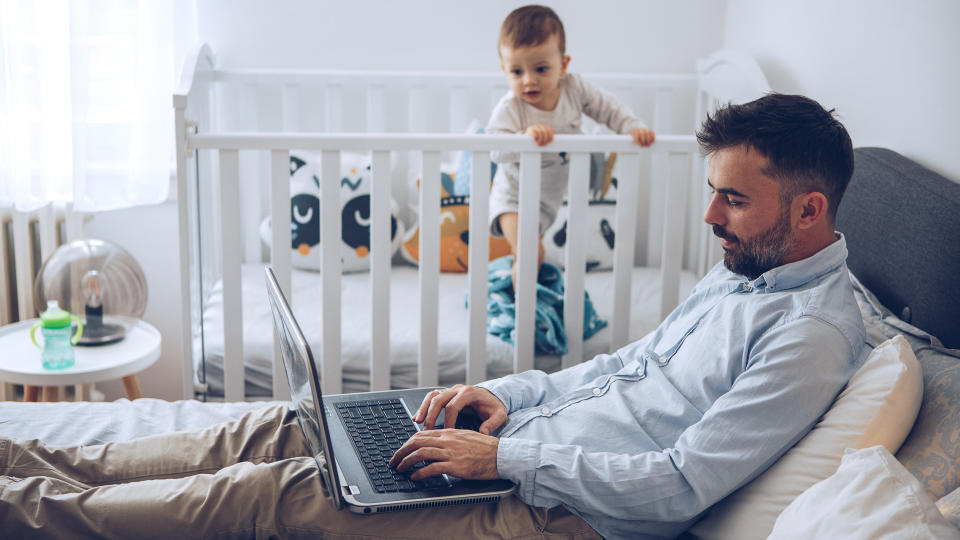 Father finishing up some work on his laptop while his baby son can't wait him to finish up and take him from crib.