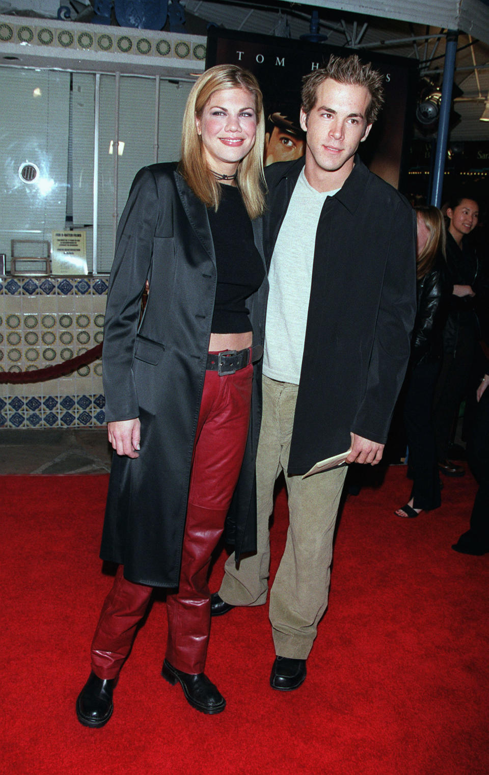 Kristen, in a black crop top, long coat, and red leather pants, with Ryan, in a casual jacket, T-shirt, and corduroys, on a red carpet